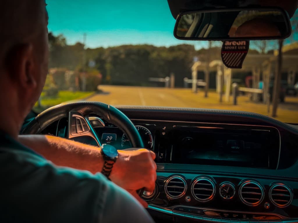 Professional chauffeur driving a Mercedes S-Class toward a sunlit arrival at a premium property