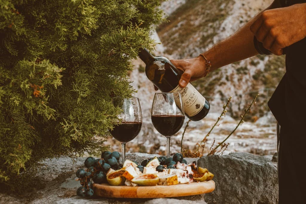 Outdoor wine tasting, red wine poured into a glass beside cheese, figs, and grapes with hills beyond